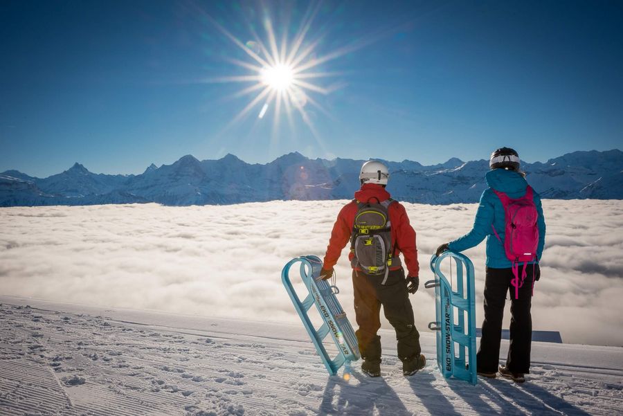 Zwei Personen im Schnee mit Schlitten blicken auf das Nebelmeer und die Berge