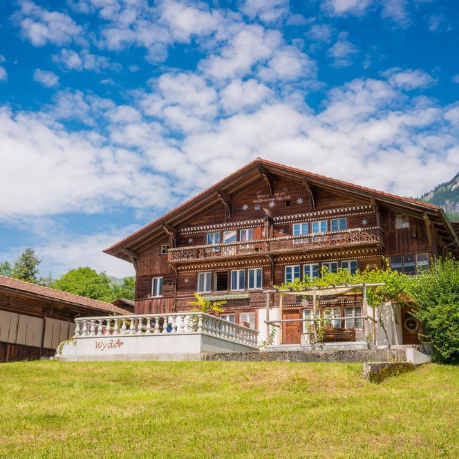Schönes Schweizer Chalet auf einer grünen Wiese in Schwanden. Der Himmel ist blau und verziert mit Schäfchenwolken.