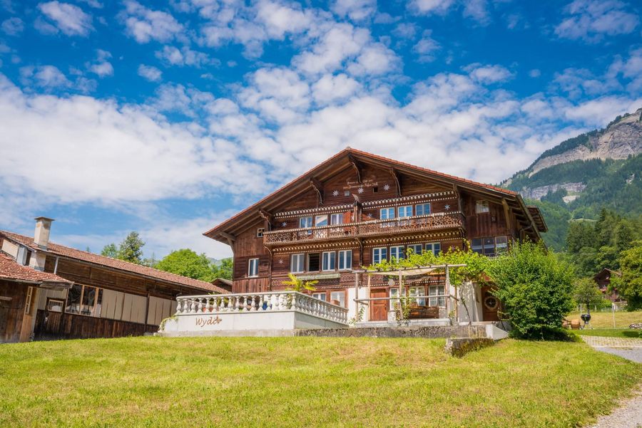 Schönes Schweizer Chalet auf einer grünen Wiese in Schwanden. Der Himmel ist blau und verziert mit Schäfchenwolken.