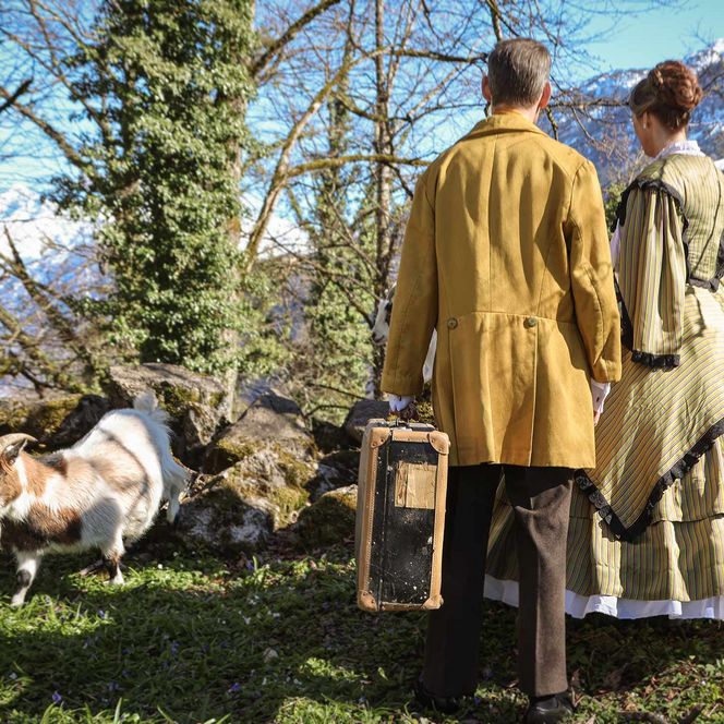 Ein Mann und eine Frau in authentischen alten Kleidern stehen, mit dem Rücken zum Betrachter neben einer weiss-braunen Ziege.