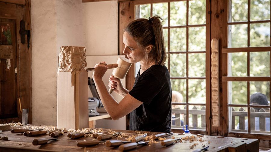 Eine Holzbildhauerin schnitzt an einem Holzstück in einem Gebäude im Freilichtmuseum Ballenberg.