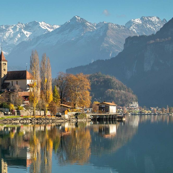 Die Kirche auf der Anhöhe in Brienz spiegelt sich im glatten Brienzersee, im Hintergrund die Berge vom Haslital.
