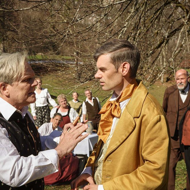 Beobachtet von weiteren Personen diskutiert ein älterer Herr mit einem jüngeren. Alle sind in authentischen älteren Kleidern zu sehen.