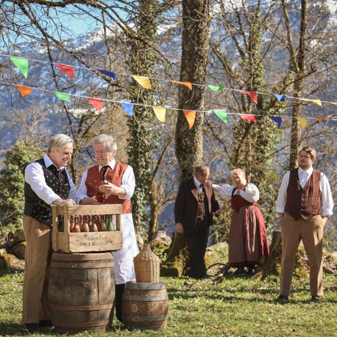 Verschiedene Darsteller in authentischen alten Kleidern stehen auf einer Lichtung. Zwei Männer hantieren mit Kisten.