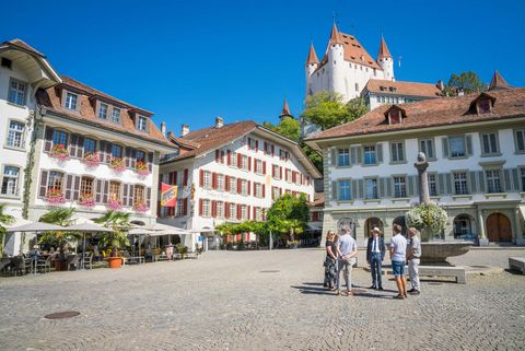 Unterwegs auf einer Themenführung hören die Gäste auf dem Rathausplatz dem Stadtführer zu. Im Hintergrund das Schloss Thun bei wolkenlosem Himmel.